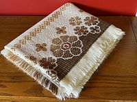 Folded vintage woven tablecloth showing floral pattern in brown, beige, and orange with fringe edges.