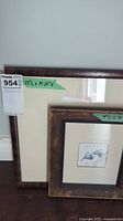 Two wooden frames against wall, one large empty frame with mat and one smaller frame with olive artwork.