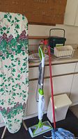 Wide view showing ironing board, Bissell steam mop, red mop, trash bin, countertop with cleaning supplies and black container box.