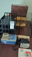 Wide photo showing office supplies including wooden letter organizer, black plastic trays, a dominoes game box, pencils, calculator, and the Pentax camera with case and manual.