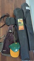 Two black fabric camping chairs in black carrying bags with Ozark Trail branding, an Oakland Athletics baseball cap, a maroon backpack, and a small black crossbody bag on wooden floor.
