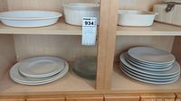 Shelf with three Corning Ware casserole dishes and multiple stacks of plates in light colors including white and gray.