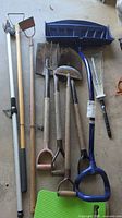 Photo showing full set of gardening tools laid on concrete floor including snow shovel, pitchforks, scrapers, rake, and shovels.