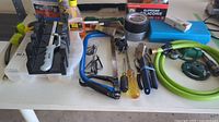 Various hand tools, including caulking gun, saw frame, utility knife, screwdriver, pliers, adhesive, tape, sandpaper and small toolbox on white table.