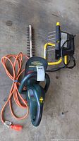 Yardworks hedge trimmer with attached orange power cord and portable yellow frame work light placed on concrete floor.