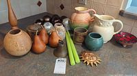 Photo showing multiple jugs in various ceramic styles and colors, several glass and metal Christmas ornaments, two green taper candles, a brass sun wall decoration, and a carved terracotta pot arranged on a counter.