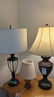 Three lamps sitting on a wooden surface including a tall metal base lamp with white fabric shade, a small wooden base lamp with beige woven fabric shade, and a medium metal base lamp with white fabric shade.