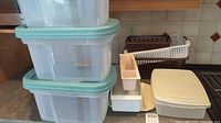 Three clear plastic storage containers with teal lids stacked on top of each other, along with white containers with off-white lids and small white inserts on kitchen counter.