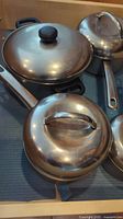 Close-up photo showing three stainless steel saucepans with domed lids, and one additional saucepan with handle visible in background.