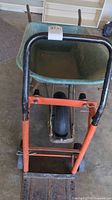Front overview showing green wheelbarrow tub on metal frame behind orange hand trolley