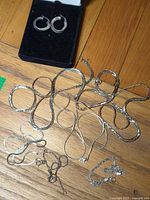 Multiple sterling silver chains and bracelets of various styles and thicknesses laid on wood surface, with earrings box in corner