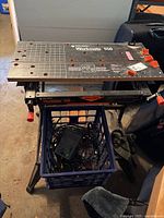 Black & Decker Workmate 550 bench showing metal top with measurement grid and orange clamps, standing on black metal legs with a milk crate below.