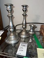 Two tall silver-toned metal candle holders with rounded bases and classic turned designs displayed side by side on a glass surface.