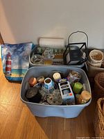 Blue plastic tub filled with assorted candles, glass candle shades, and candle holders sitting on wooden floor next to woven baskets and holiday bag with lighthouse image