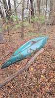 Green canoe laying upside down on leaf-covered ground and surrounded by trees and branches, with signs of outdoor storage.