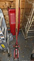 Red metal car ramp and red 2 ton hydraulic floor jack stored upright indoors on concrete floor with wood wall background, showing debris and rodent evidence.