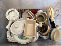 Top view of bin containing white handled pots with lids, plates, crocks, and bowls stacked inside the bin