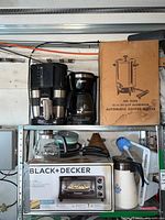 Shelf with boxed Black & Decker toaster oven, boxed Automatic Coffee Maker No. 9308, Black & Decker coffee maker, Gevalia glass coffee carafe, and some cups and pans.