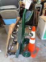 Photo showing a green metal dolly with wheels alongside a wood dolly and an orange traffic cone in a garage environment.