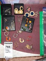 Top-down view of multiple assorted earrings laid out on purple cloth, showing various styles including hoops, clip-ons, drop earrings, and studs.