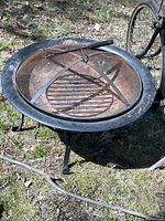 Full view of round outdoor fire pit on grass, showing bowl, legs, mesh screen, grill grate, and fire poker tool.