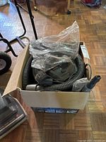 Photo showing the ash vacuum main unit inside a cardboard box along with the flexible hose and nozzle attachments wrapped in plastic.