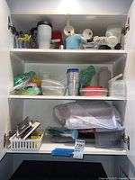 Three-shelf kitchen cabinet with various kitchen gadgets and food storage containers including thermoses, plastic containers with lids, measuring cups, and ice trays