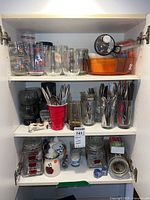 Shelf with various glassware including drinking glasses with vintage prints and more clear glasses, flatware sets in cups and tins, black coffee grinder, orange baking dish, soap dispenser, jars and bar accessories.