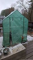 Frontal view of the outdoor greenhouse with green translucent cover and black metal frame on a wooden deck, with flower pots visible inside.