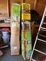 Two long cardboard boxes leaning against shed wall: one yellow Spring Gardener box, one plain green/brown box.