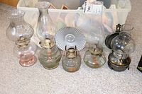Five glass oil hurricane lamps arranged on carpet, showing varied chimney and base shapes, metal burners visible