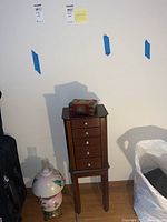 Full view of wood standing jewelry chest with small carved box on top and surrounding items (lamp and bag).