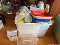 White plastic bin filled with assorted plastic food storage containers and lids in various sizes and colors including red, green, blue lids.