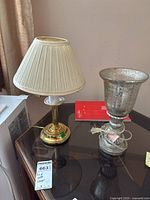 Two table lamps displayed on a glass tabletop. One lamp has a gold-tone metal base with a cream fabric pleated shade; the other features a silver base with a textured glass shade. Cords visible and tag present on one.