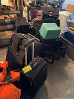 Multiple pieces of luggage including rolling suitcases and travel bags stacked on garage floor with one plastic storage bin on top.