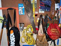 Photo showing wall pegboard holding various tools including a large saw, yellow extension cord coiled, leather tool belt, and hand pruner type cutter.
