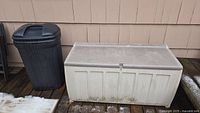 Beige resin outdoor storage chest and black plastic barrel bin side by side showing dirt and weathering on the storage chest.