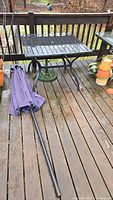 Black metal square outdoor table with a slatted top and folding legs, accompanied by a collapsed purple fabric umbrella and a green metal umbrella stand with decorative pattern.