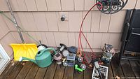 Wide shot showing all items grouped on wooden deck against siding. Includes solar lights, watering can, owl statue, snow shovel, plant stand, basket.