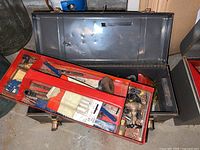 Open metal tool box with red removable trays showing a brass valve, masonry drill set, manual drill and various small hand tools inside.