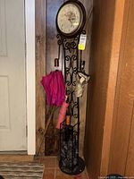 Front view of black metal umbrella stand with mounted clock at top. Three umbrellas hang from the stand. One bright pink, one light pink, and one beige. Blue bag placed on floor next to stand.