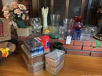 Wide view of multiple glass vases and ceramic vases on hearth with several metal decorative tins stacked on floor in front and to side.