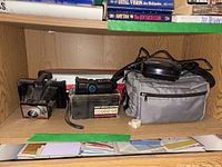 Shelf containing vintage cameras, a camera lens box labeled 'Underground Macro Zoom Lens 80-200mm', gray camera bag, and cables.