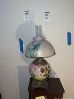 Full view of vintage glass and brass floral table lamp on wood surface, showing glass globe body and matching glass shade with chimney, decorated with hand-painted floral motif.