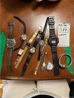 Top-down photo of nine watches arranged on a wooden surface, showing variety of styles including metal, leather, and plastic bands.