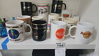 Photo of assorted advertising and souvenir coffee mugs from Universal Pictures, Hard Rock Cafe, Chambers Development, UPS, Yellow Freight System, and more, arranged on a white shelf.