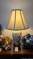Full view of the Asian ceramic base table lamp lit, showing the fabric shade, engraved floral designs on ceramic, wood pedestal base, and overall lamp proportions