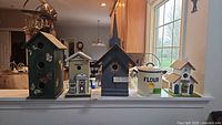 Five decorative birdhouses and one ceramic flour canister arranged on a ledge, showing variety in shapes, sizes, and decorations including painted butterflies and a chapel motif.