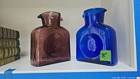 Front and side views of purple and blue Blenko glass decanters on a white shelf with an 8 inch label on the blue decanter.
