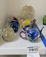 Front view of nine hand blown glass paperweights including fish, bird, whale, teapot with flower, and frosted dog on a white shelf.